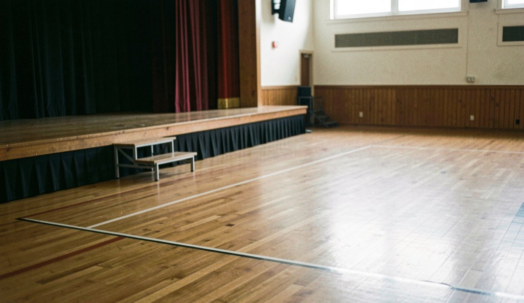 An empty hardwood ballroom floor is viewed from the side with a small stage and clear walkways.